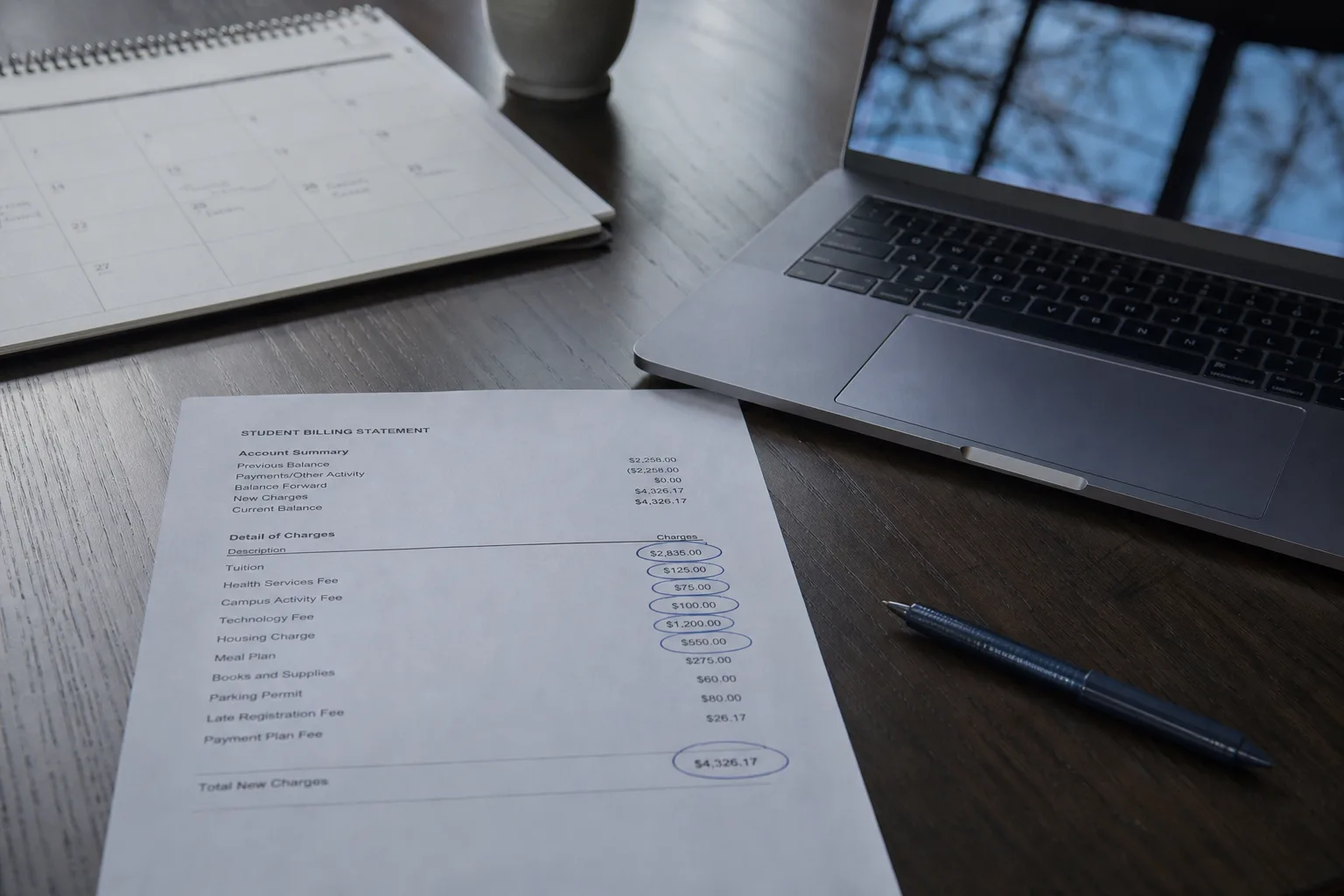 A student billing statement on a kitchen table beside a laptop, a calendar, and a pen, with several line items circled