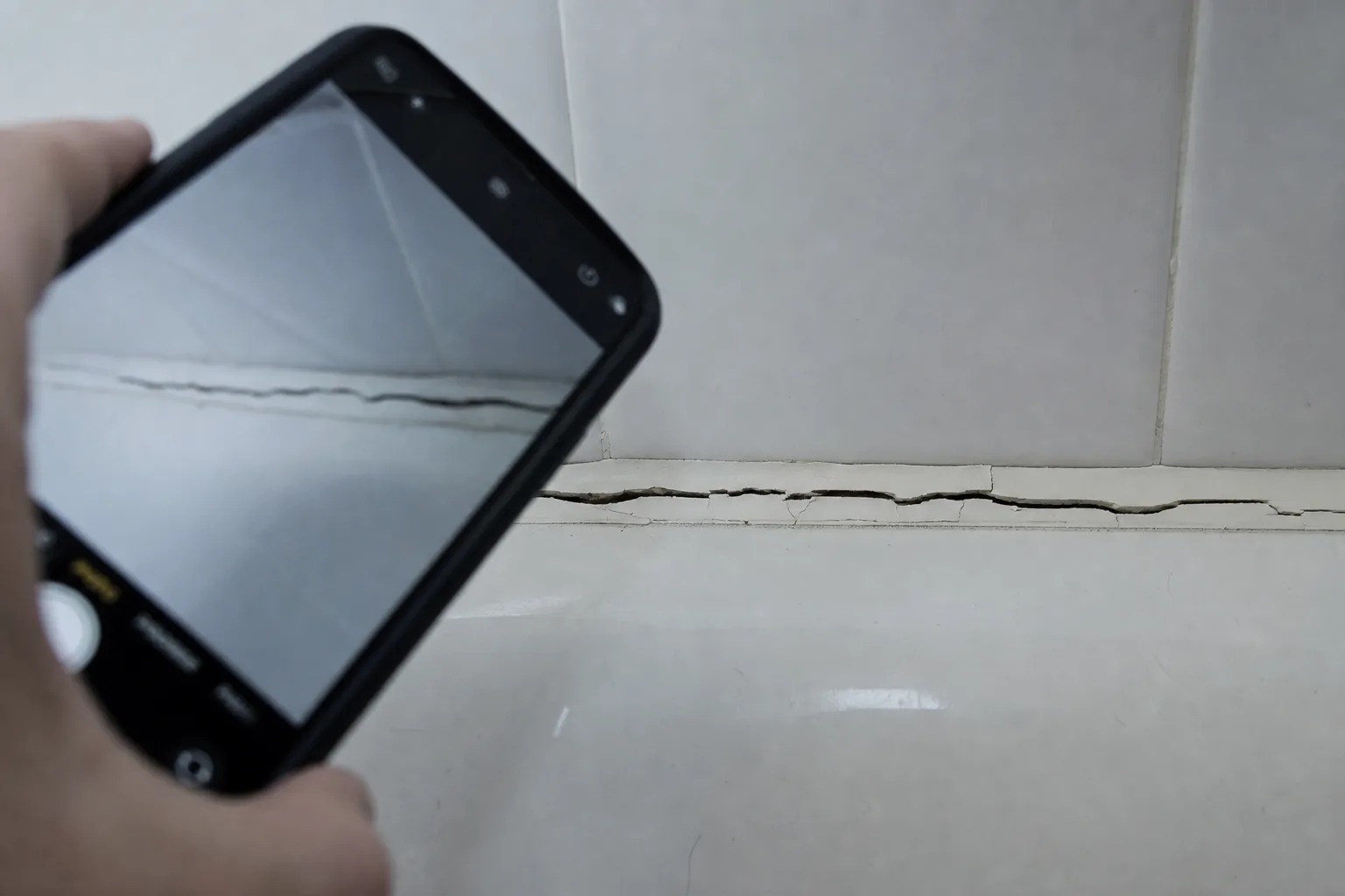 Close-up of a cracked, separating bead of white caulk where a bathtub meets a tiled shower surround in an apartment bathroom, with a smartphone held up photo...