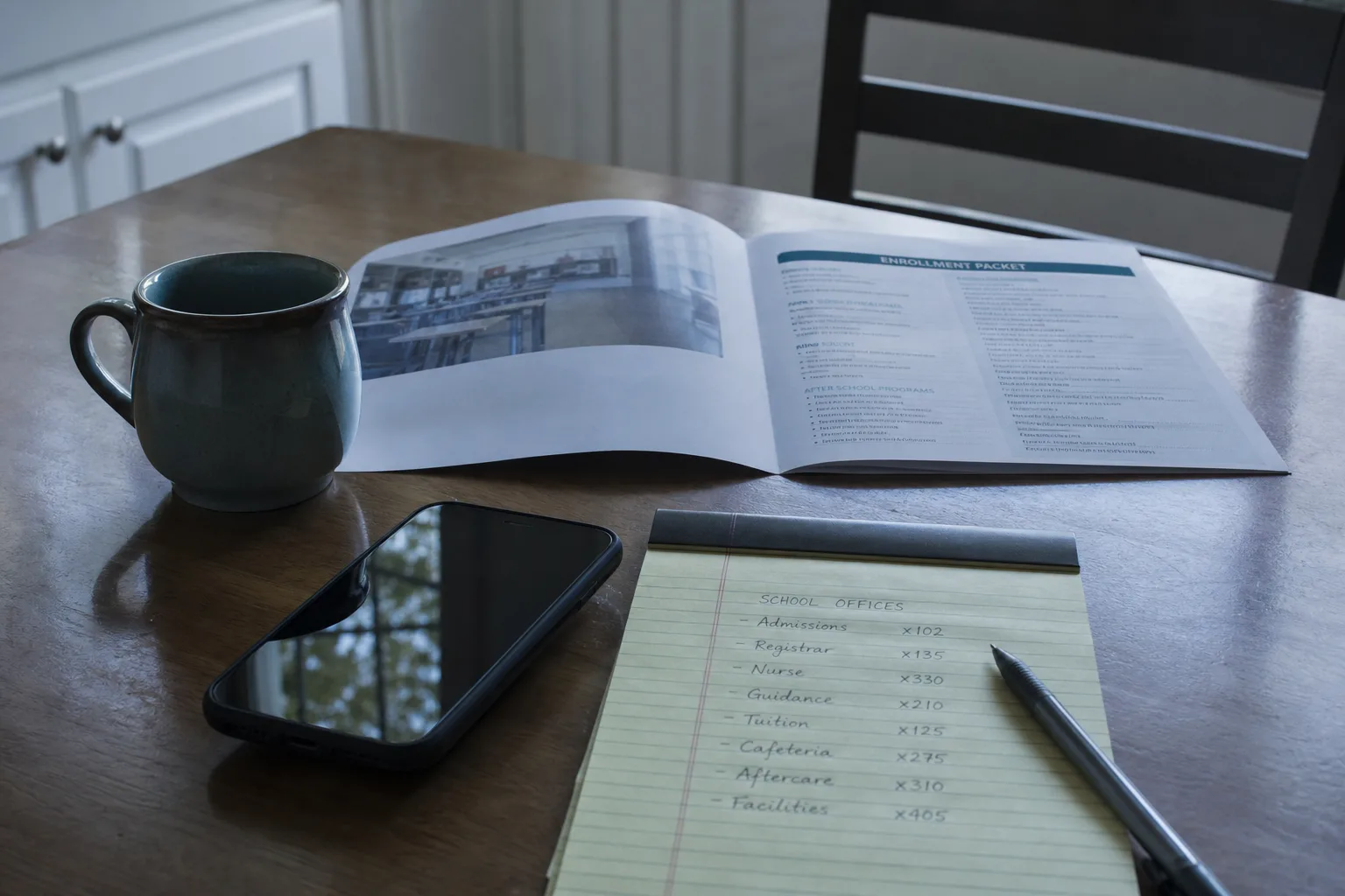 A kitchen table with a school enrollment packet open, a handwritten list of office names and phone extensions on a legal pad, and a phone face up beside a co...