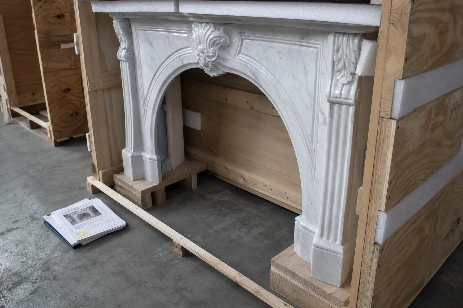 A carved marble fireplace mantel resting on wooden blocking inside a partly assembled plywood shipping crate, with foam padding and a stack of labeled paperw...