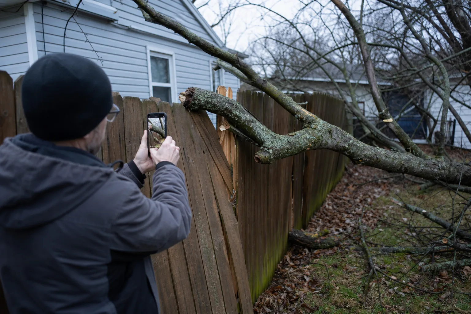 A residential property line where a fallen tree limb has damaged a wooden fence between two suburban back yards, with a homeowner photographing the damage on...